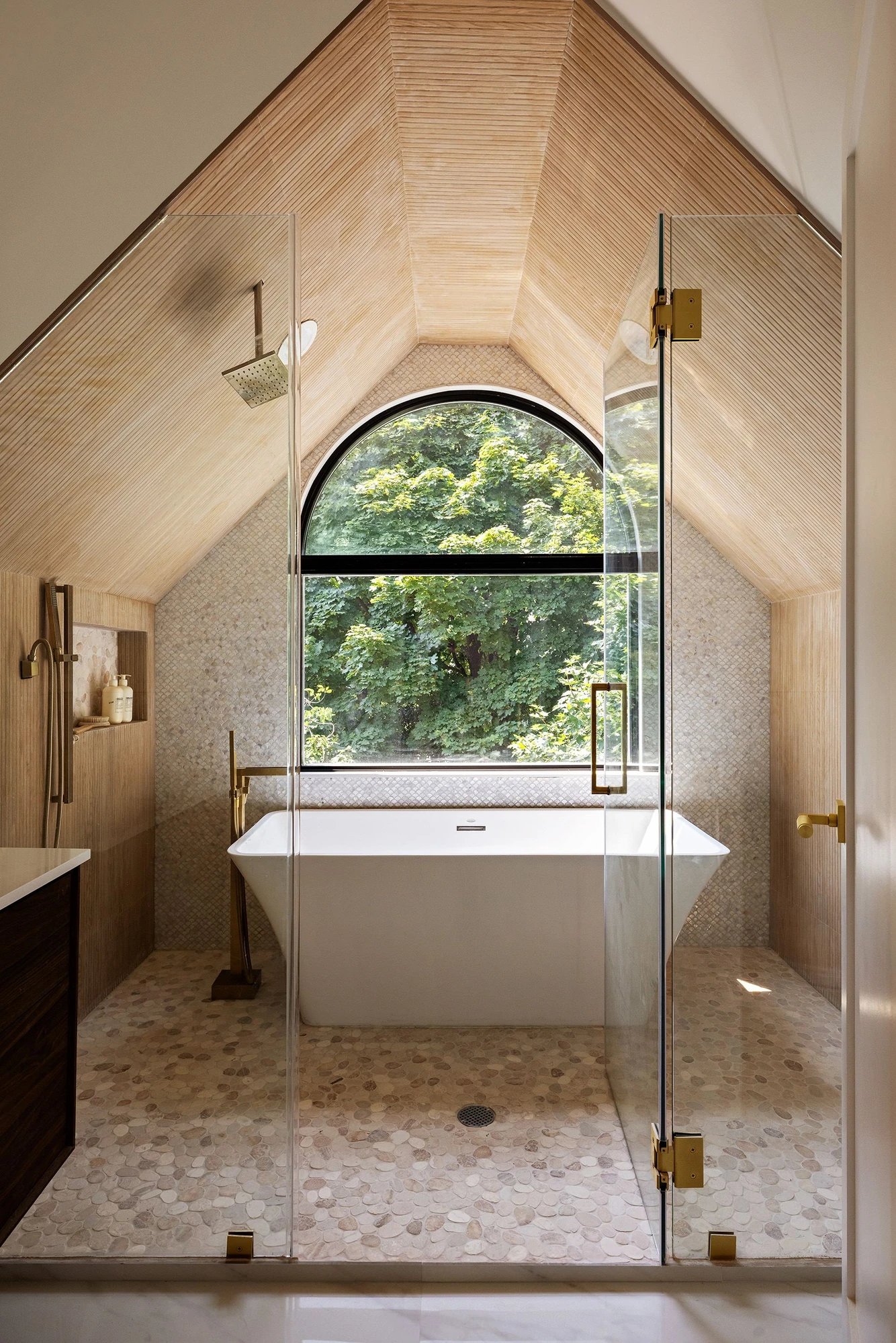 Treetop wet room with a vaulted wood ceiling, arched window, pebble floor, and freestanding tub at 534 East 24th Street