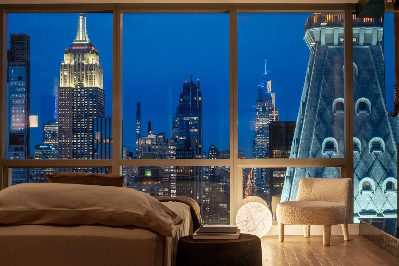 Bedroom at night with the illuminated Empire State Building and MetLife Clock Tower beyond floor-to-ceiling windows, beside a bed and rounded chair