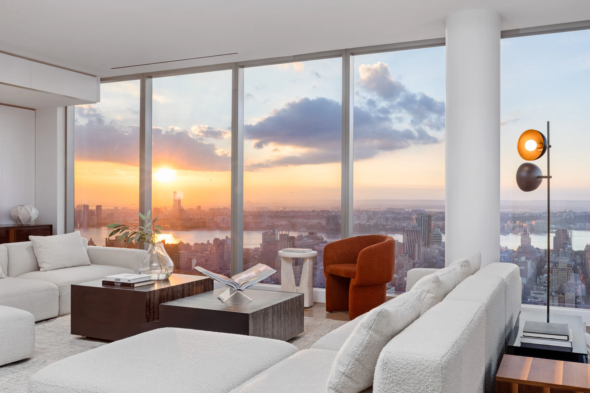 Sunset over the river beyond the living-room windows, with pale sofas and a rust-colored chair in the foreground