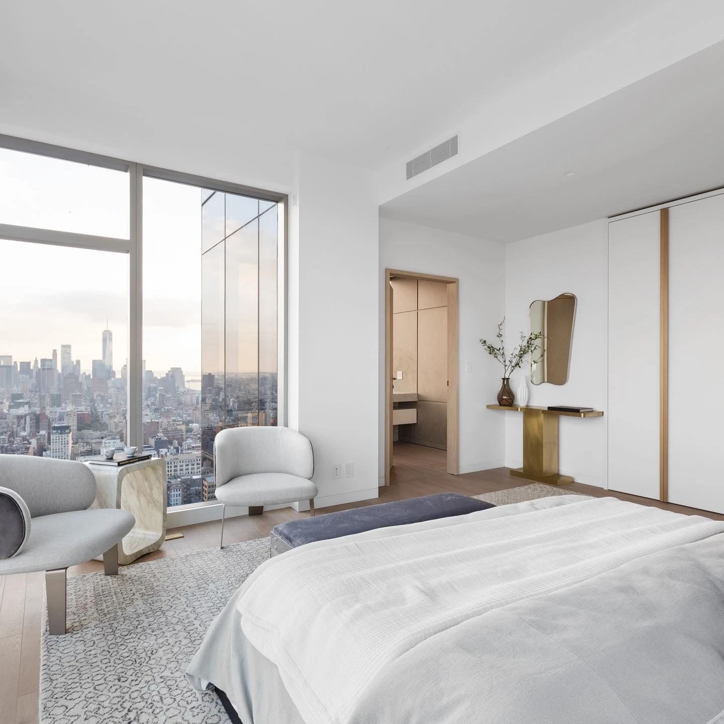 Bedroom with pale bedding, two chairs by tall windows, and the downtown Manhattan skyline in the distance