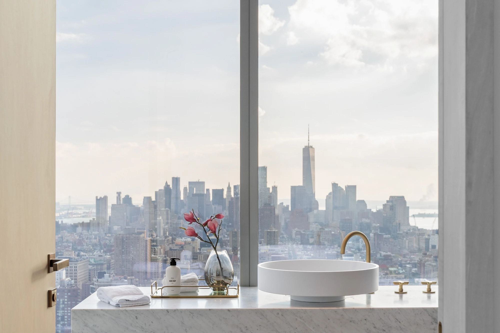 Round vessel sink and gold-toned faucet in front of a bathroom window framing downtown Manhattan and One World Trade Center