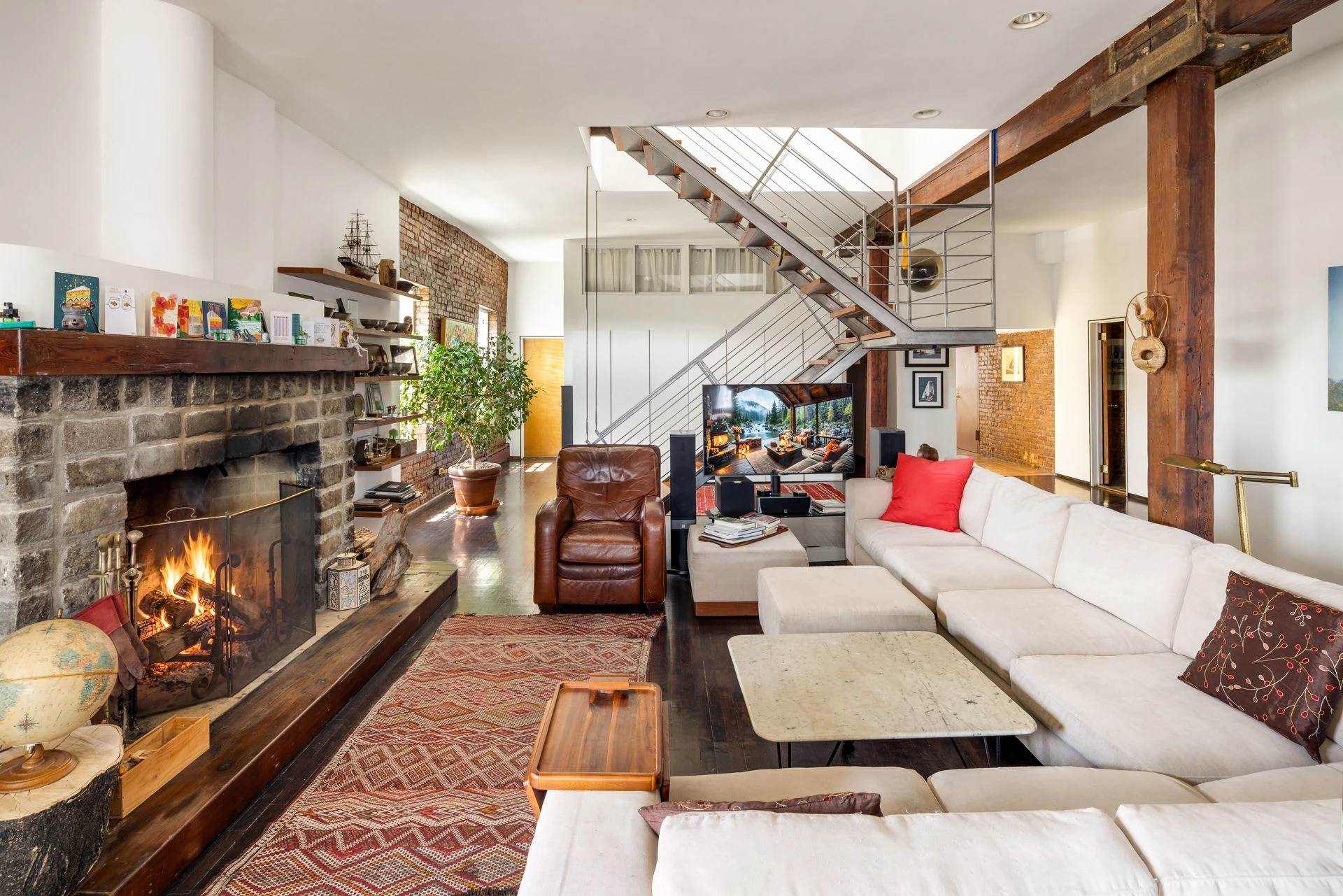 Living room with a broad stone fireplace, pale sectional sofa, leather chair, brick walls, exposed beams, and a staircase in the background