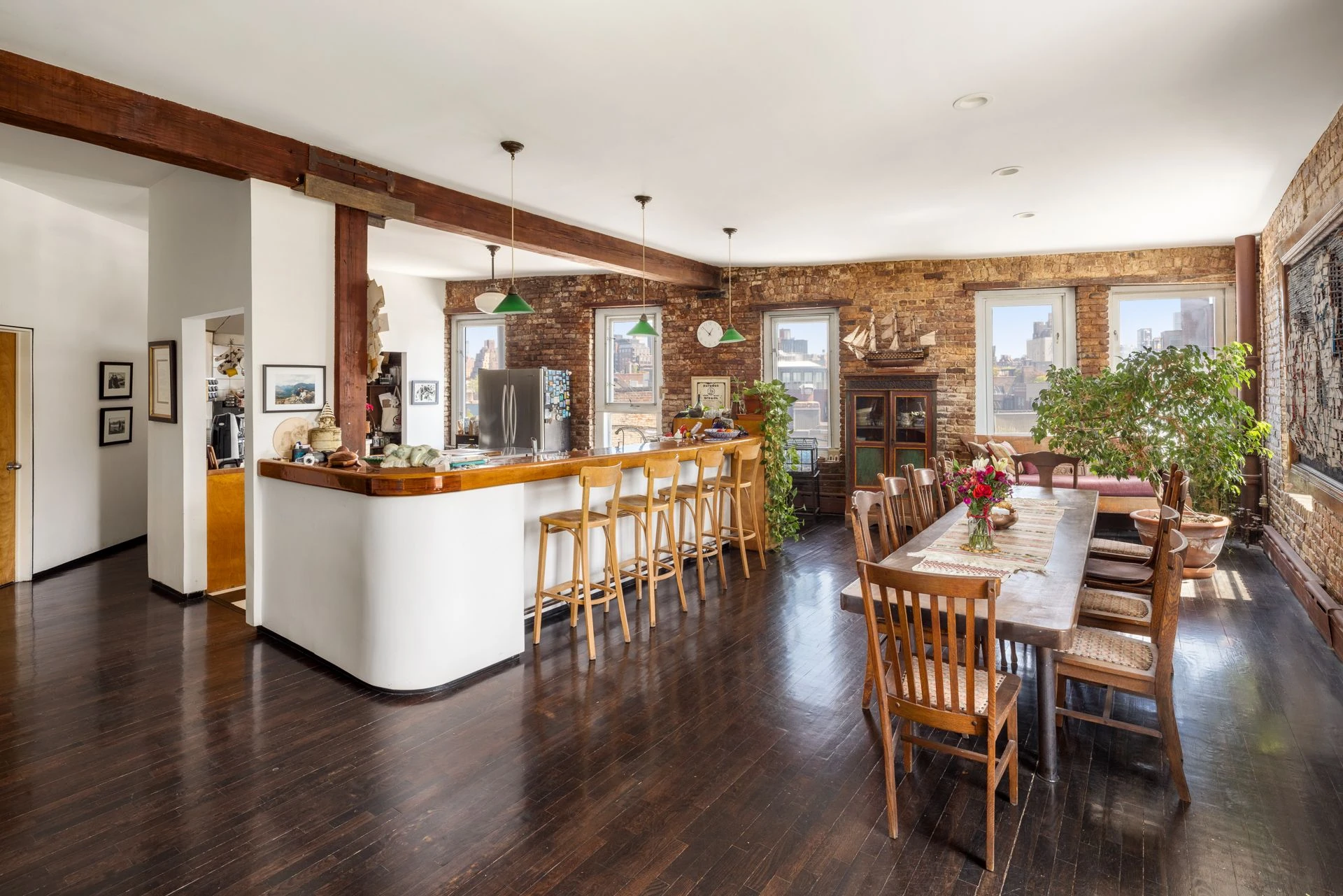 Curved wood breakfast counter with stools and green pendant lights beside a dining table, brick window wall, and exposed wood beams