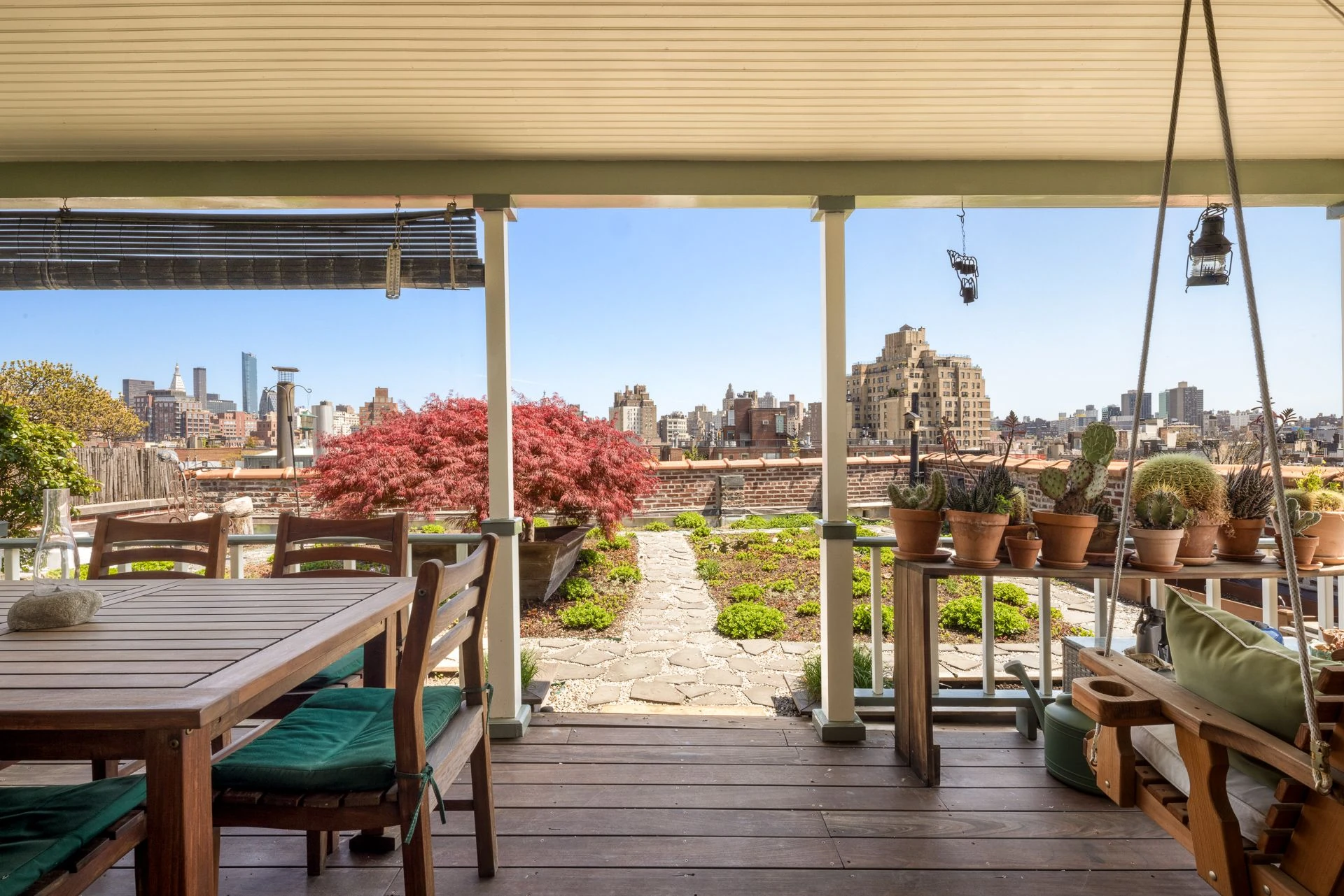 Rooftop garden seen from beneath the white porch ceiling, with a wood table, swing, planted terrace, and city buildings beyond