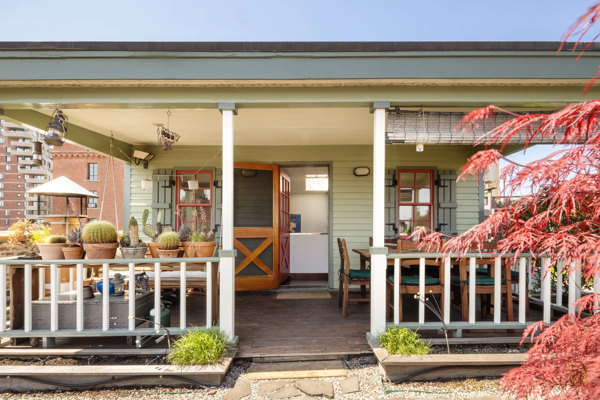 Pale green cottage with a wood screen door, shutters, white porch posts, potted cacti, a swing, and an outdoor dining table
