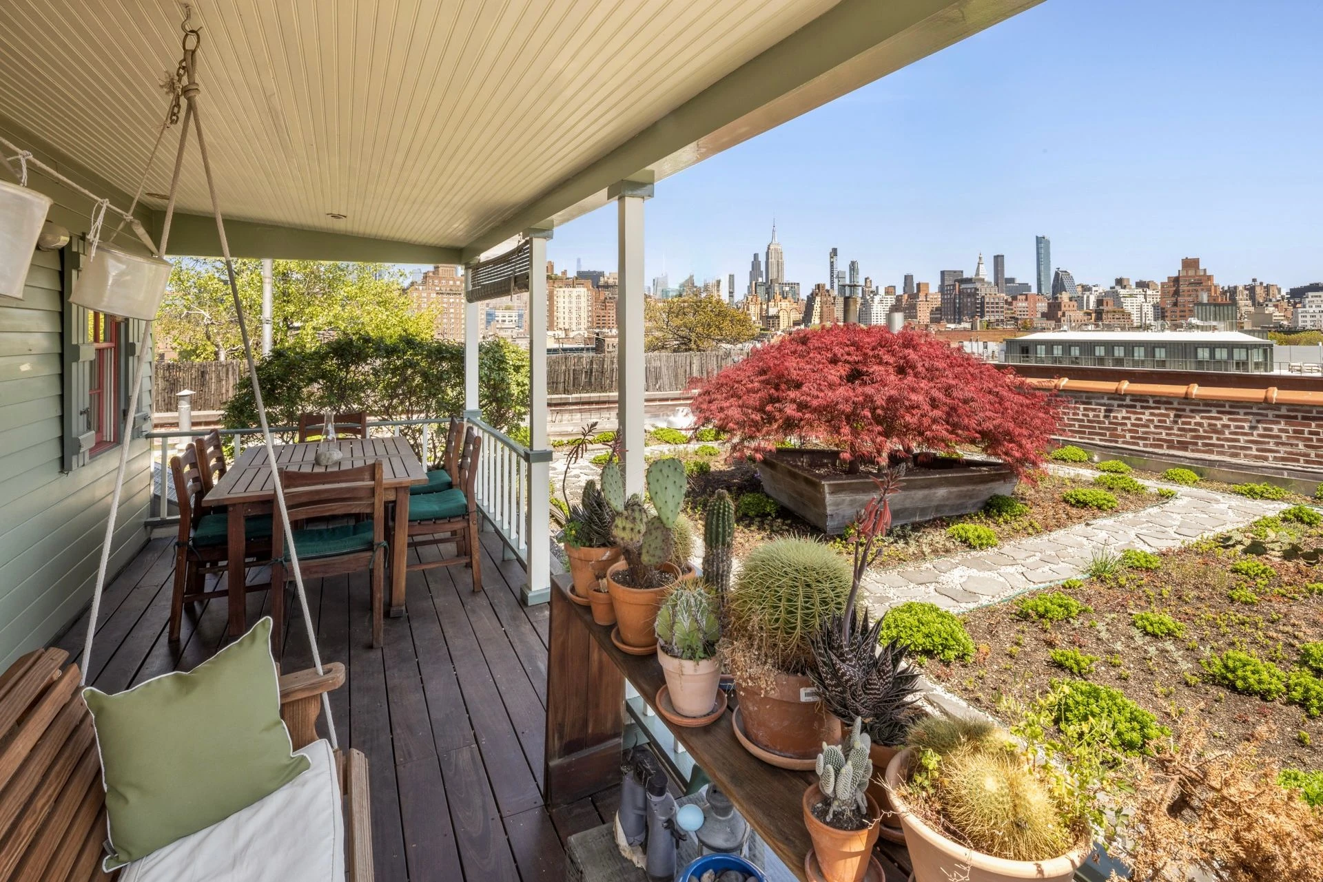 View along the covered rooftop porch, with a swing, dining table, potted plants, a red-leafed tree, and the Empire State Building beyond